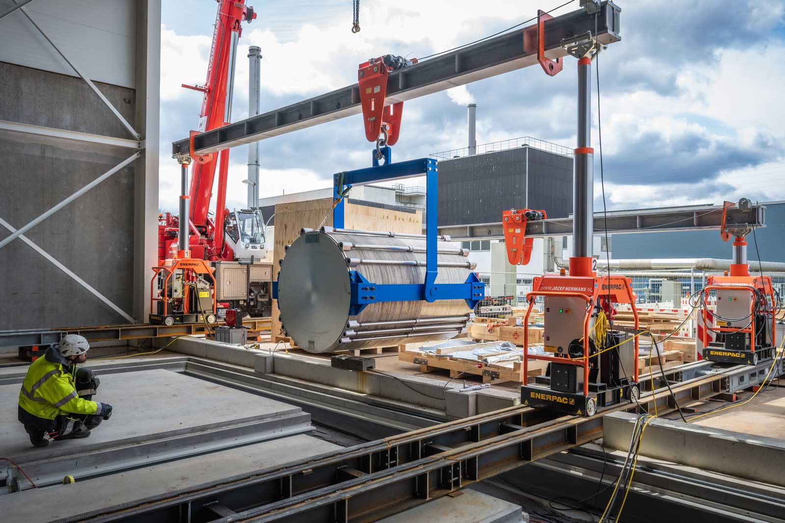 Eine Baustelle mit einem Kran, der ein großes zylindrisches Objekt hebt. Arbeiter und Maschinen sind sichtbar.