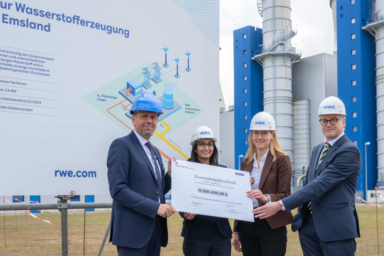 Gruppenfoto vor einem Plakat zur Wasserstofferzeugung mit einem Scheck über 8.000.000,00 Euro und Industrieanlagen im Hintergrund.