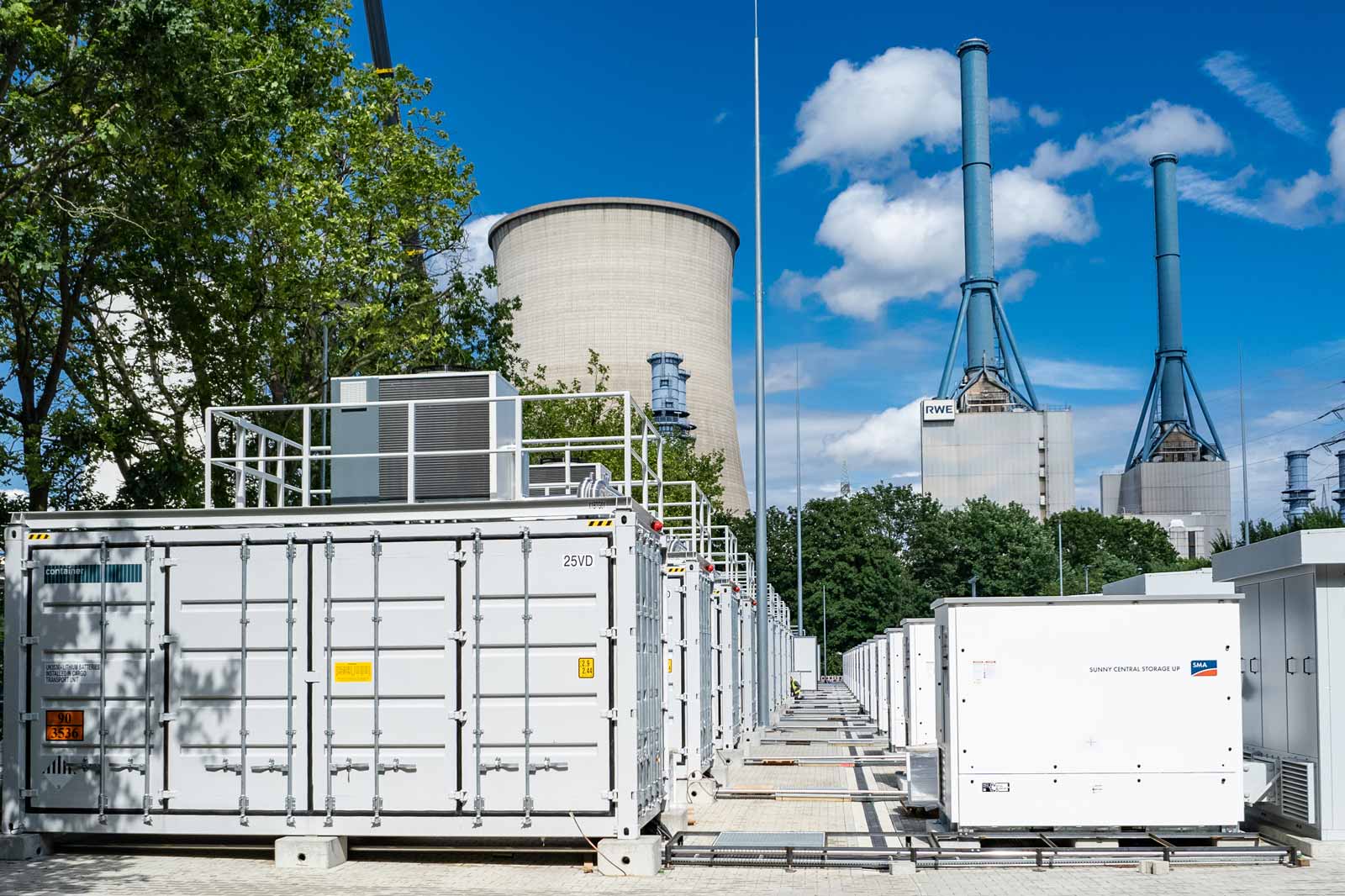 Eine Reihe von Containern zur Energieerzeugung vor einem Kühlturm und weiteren Industriegebäuden an einem sonnigen Tag.