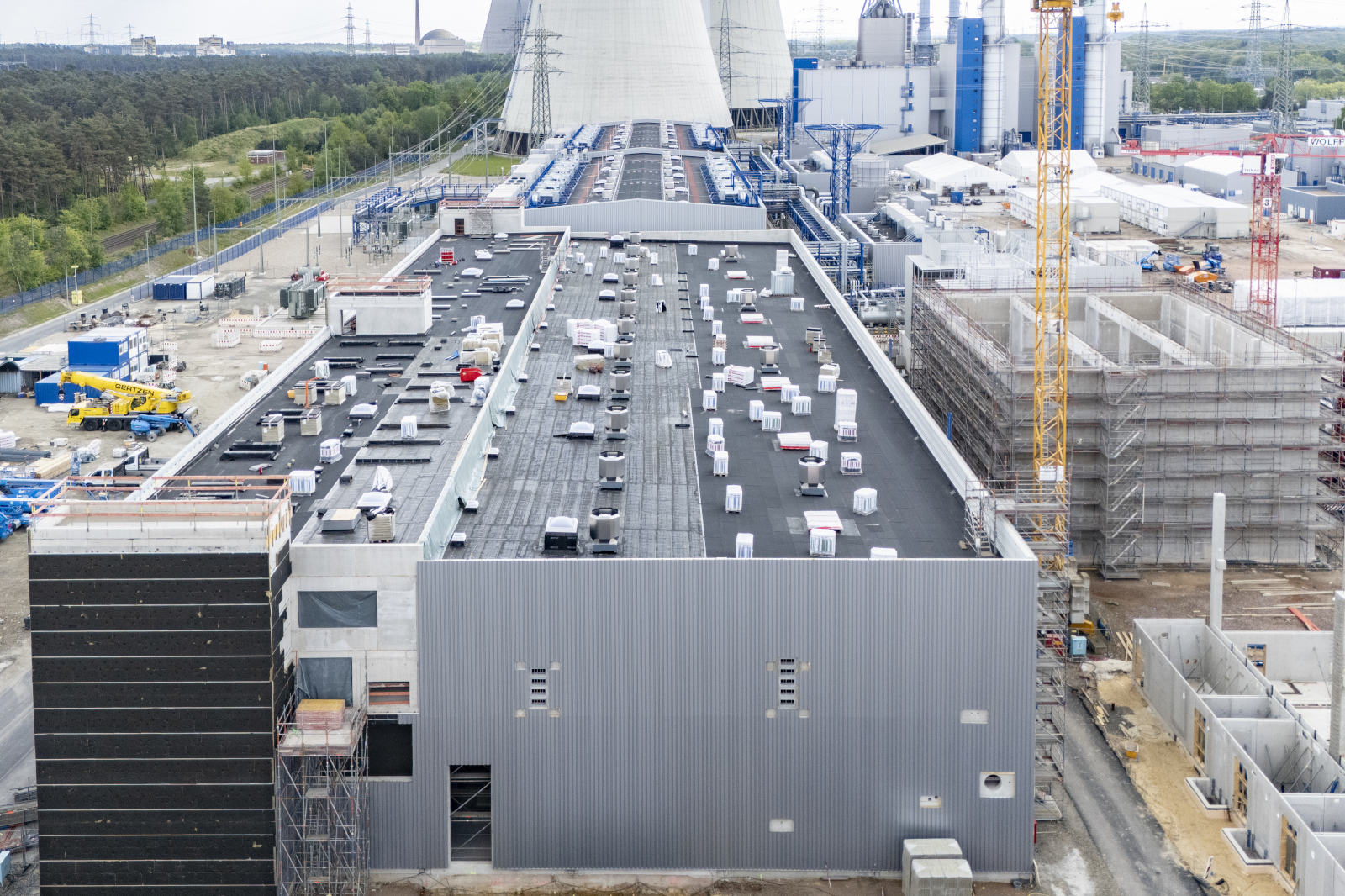 Ein großes rechteckiges Industriegebäude mit grauer Wellblechfassade und schwarzem Flachdach, auf dem mehrere Lüftungsgeräte stehen; Kräne und Kühltürme sind im Hintergrund sichtbar.