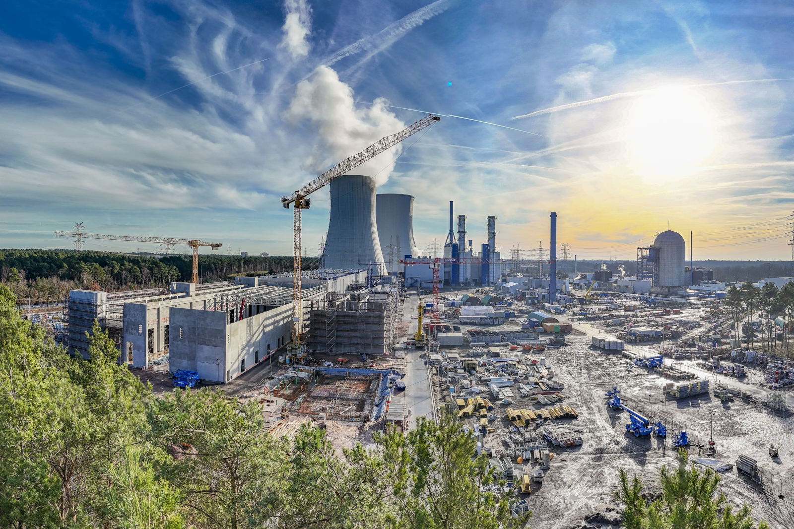 Eine weitläufige Industriebaustelle mit zwei massiven Betonkühltürmen und hohen Kränen vor einem klaren blauen Himmel mit ziehenden weißen Wolken.