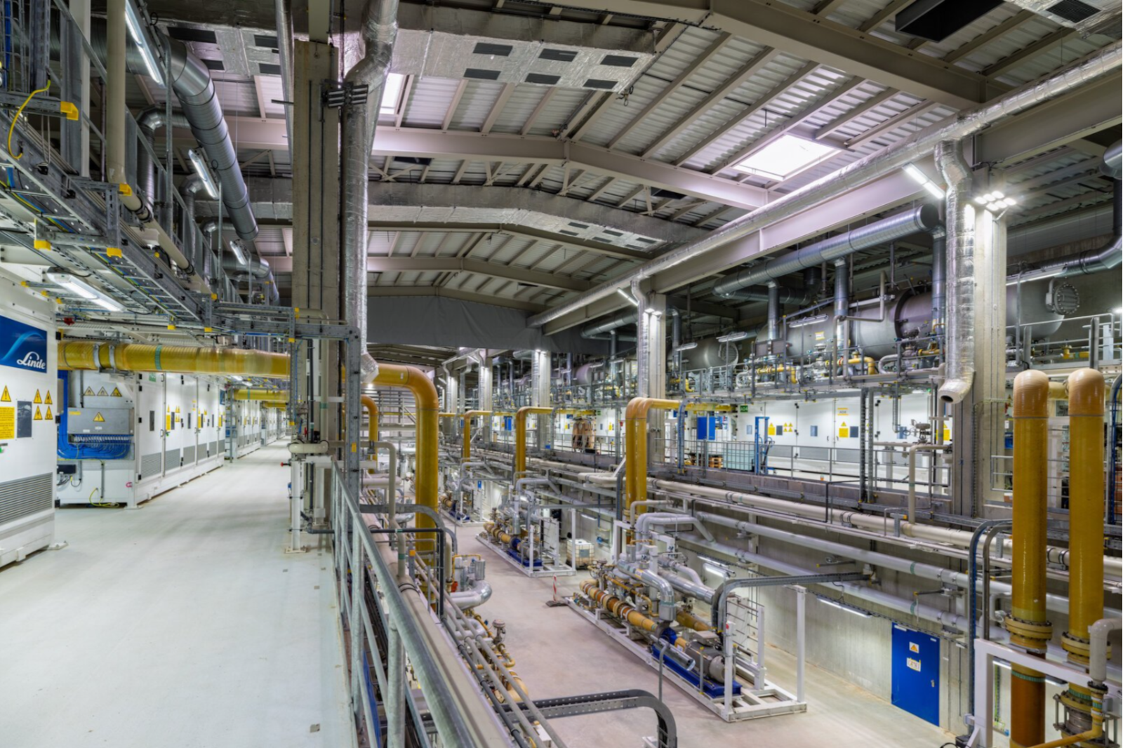 Große Industriehalle mit einem Geflecht aus silbernen Metallrohren und gelben isolierten senkrechten Leitungen unter einem hohen gewölbten Dach mit Oberlichtern.