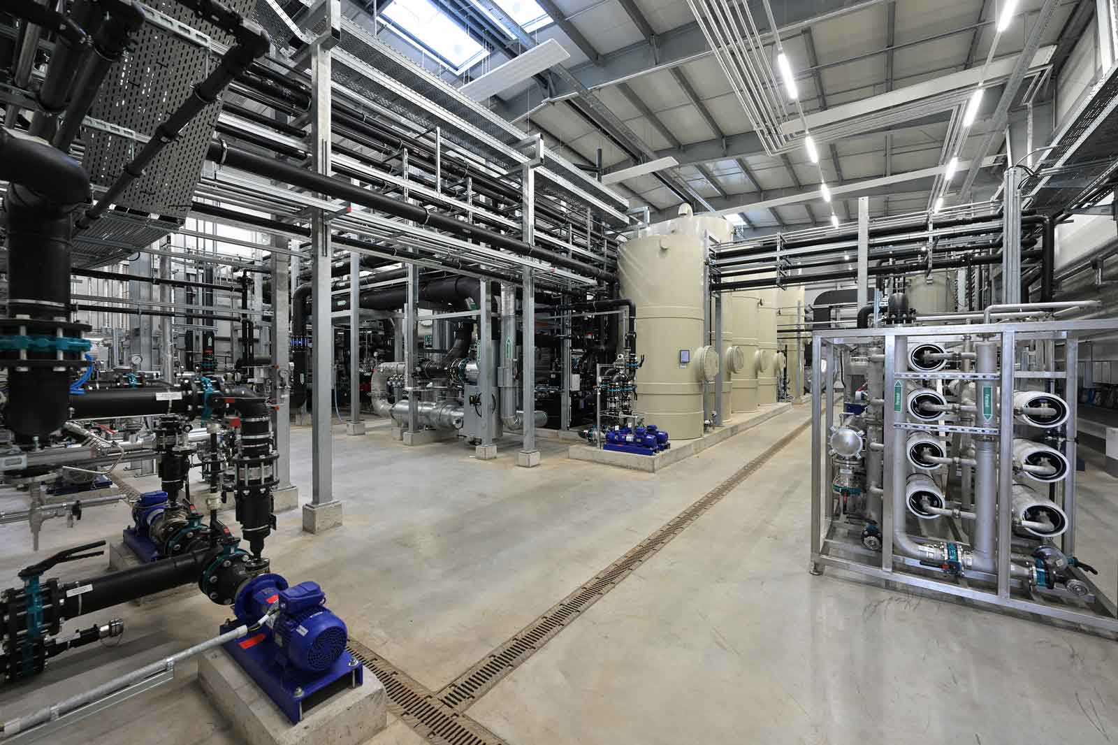 Eine geräumige industrielle Anlage mit zahlreichen Rohren, Tanks und Pumpen, die ordentlich unter einer hellen Decke angeordnet sind.