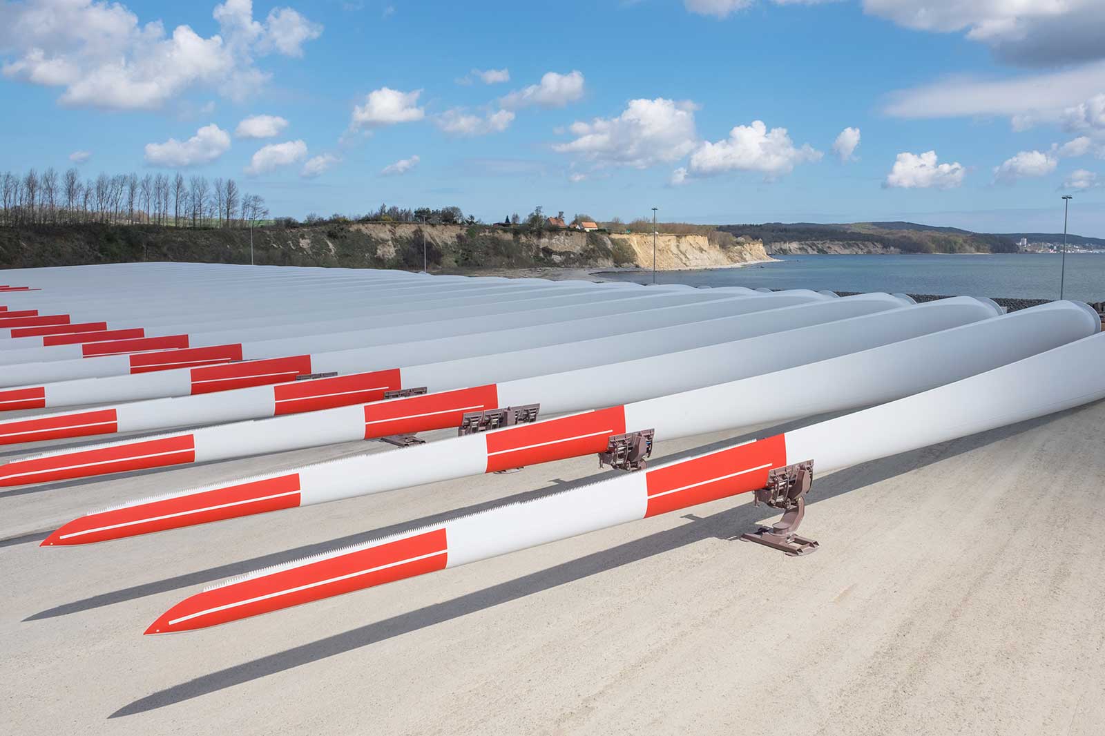 A row of wind turbine blades with red and white stripes, resting on a concrete surface near the coast.