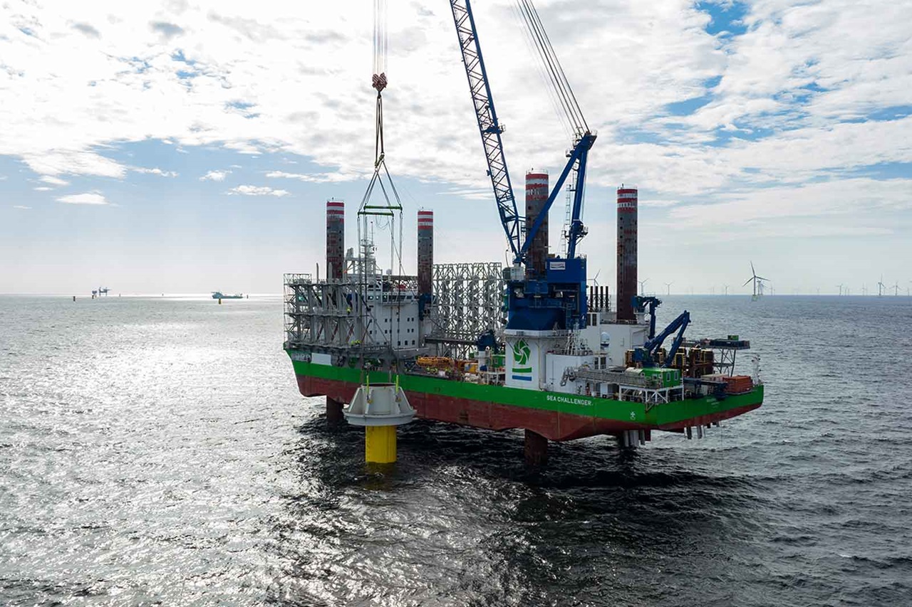 An offshore ship with a large crane lifts a yellow structure from the water, surrounded by wind turbines in the background.