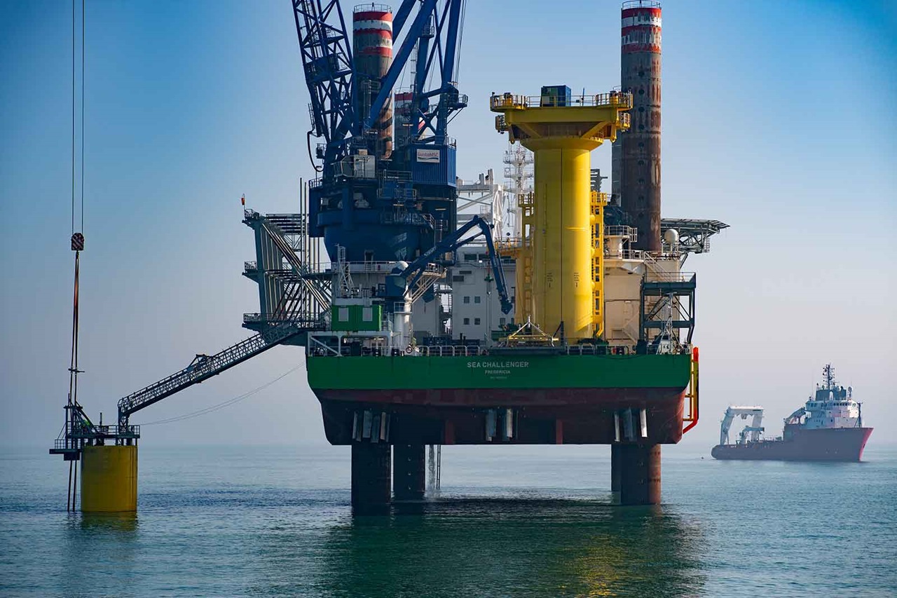An offshore drilling rig stands in the water, featuring a yellow tower and a ship in the background.