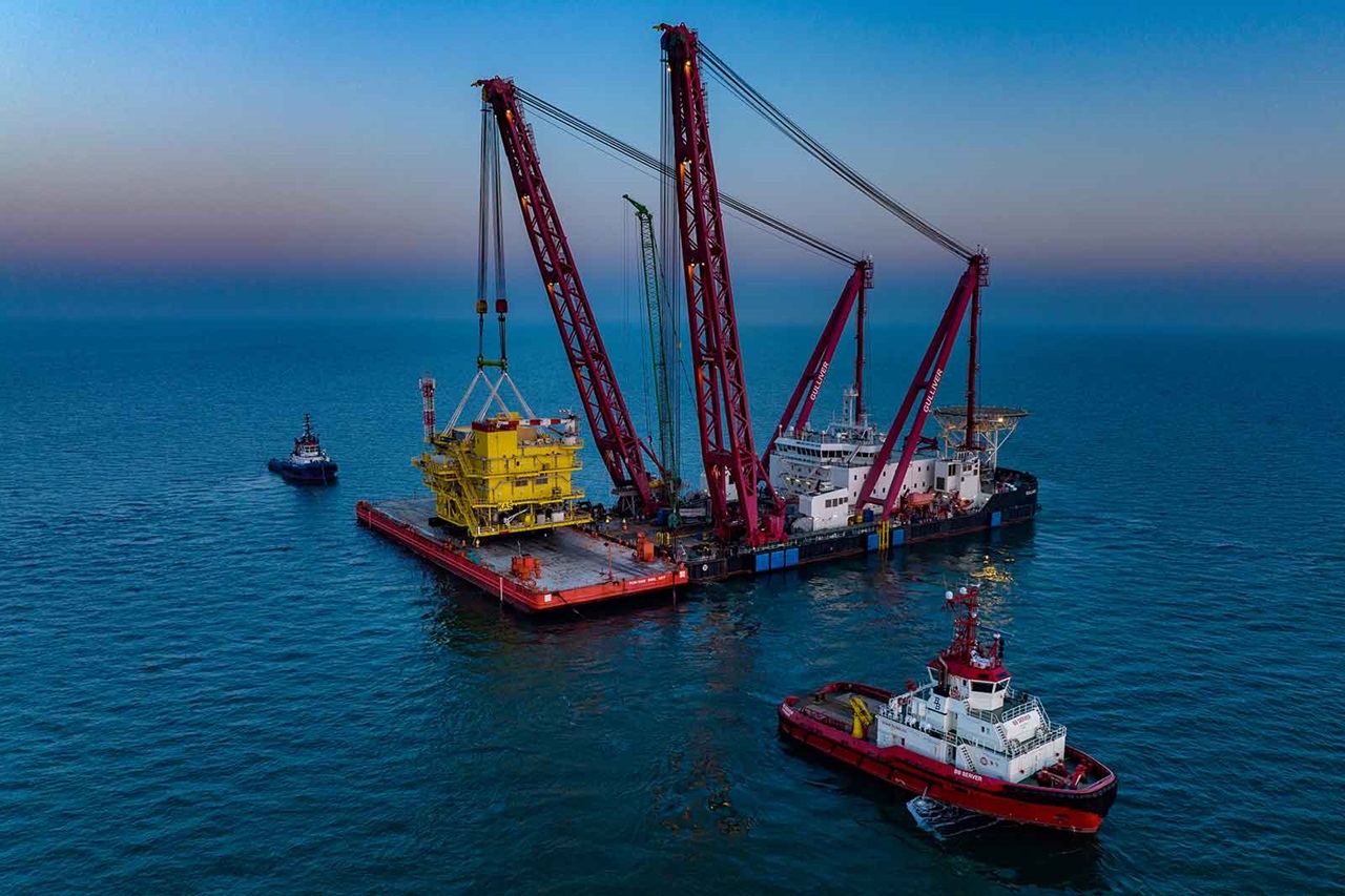 An impressive offshore vessel lifts a yellow platform from the water. Two tugboats are nearby.