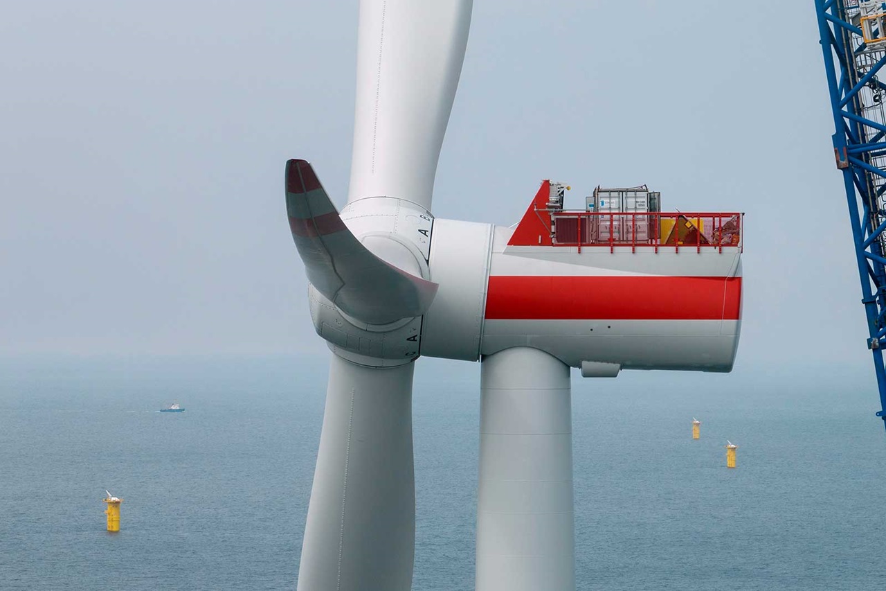 A wind turbine with a large rotor and a red platform section, set above the sea with yellow buoys in the background.