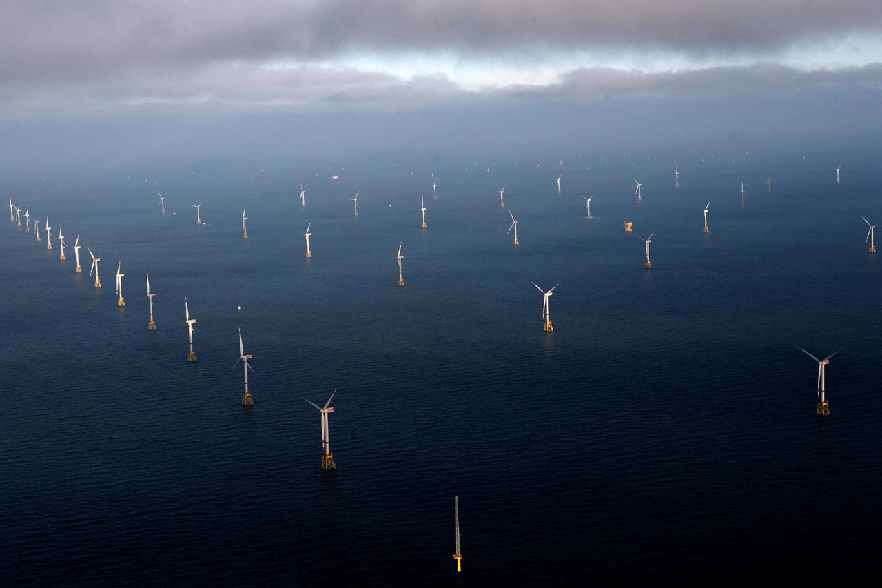 Ein Luftbild zeigt viele Windkraftanlagen im Meer, umgeben von Wasser und einem bewölkten Himmel.