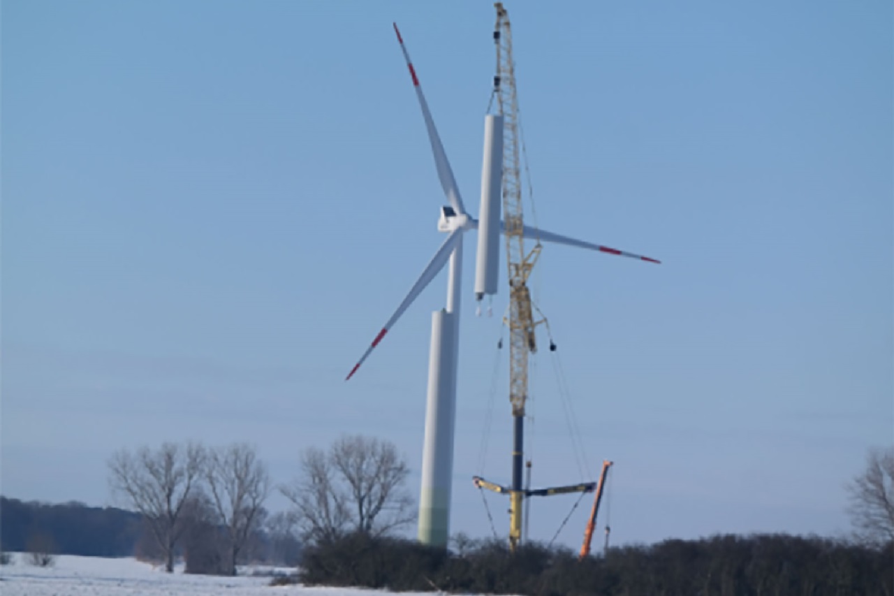 A wind turbine is being assembled by a crane in the snow, surrounded by trees and clear sky.