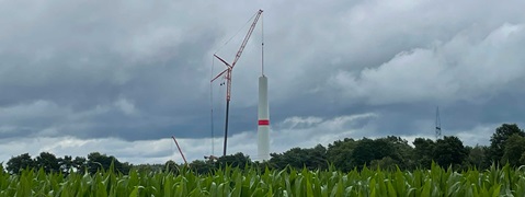 Construction of a wind turbine in the background with a crane, surrounded by a green field and cloudy sky.