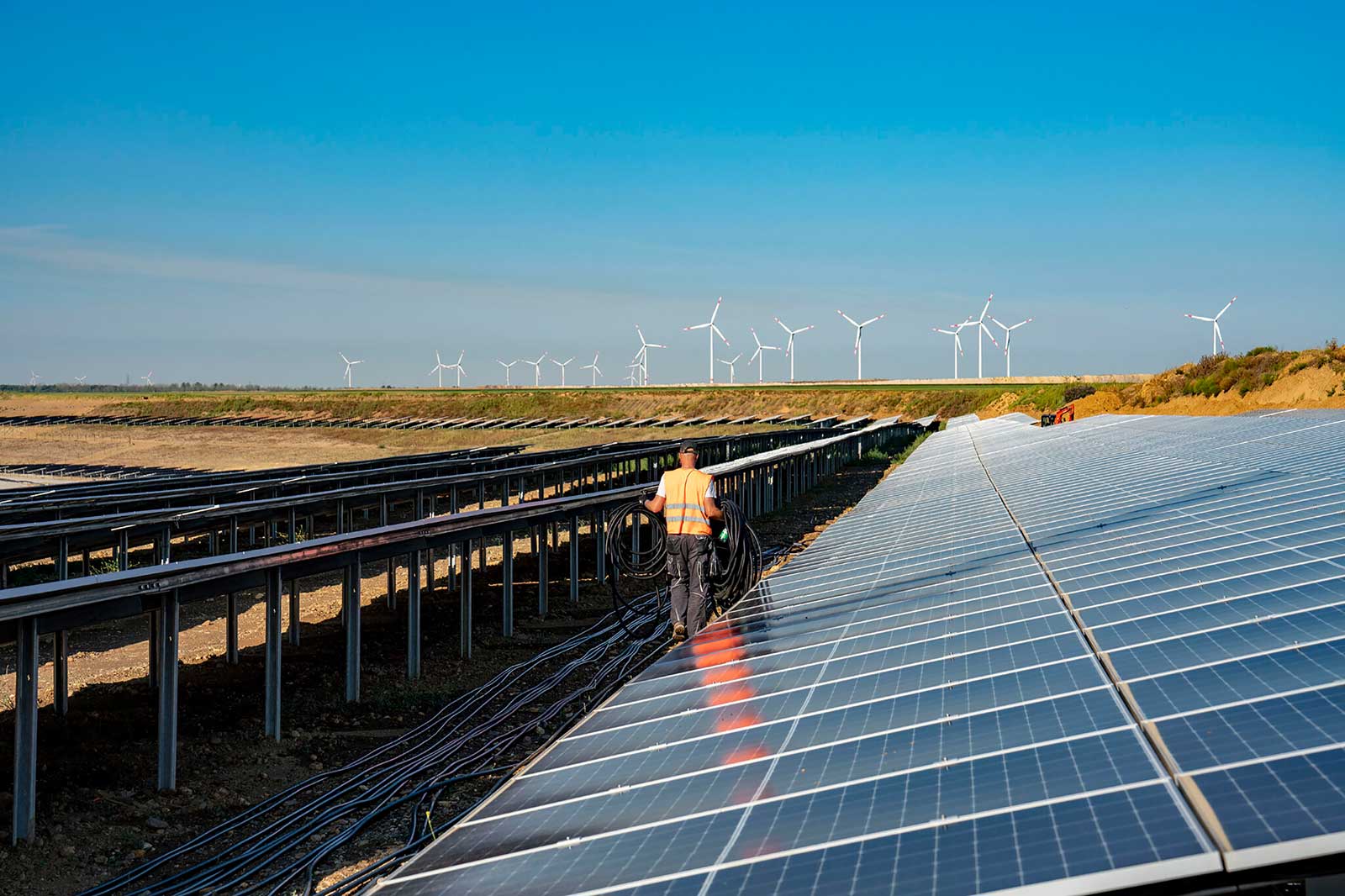 Ein Arbeiter auf einem Solarfeld trägt Kabel zwischen Reihen von Solarpanels, mit Windkraftanlagen im Hintergrund.