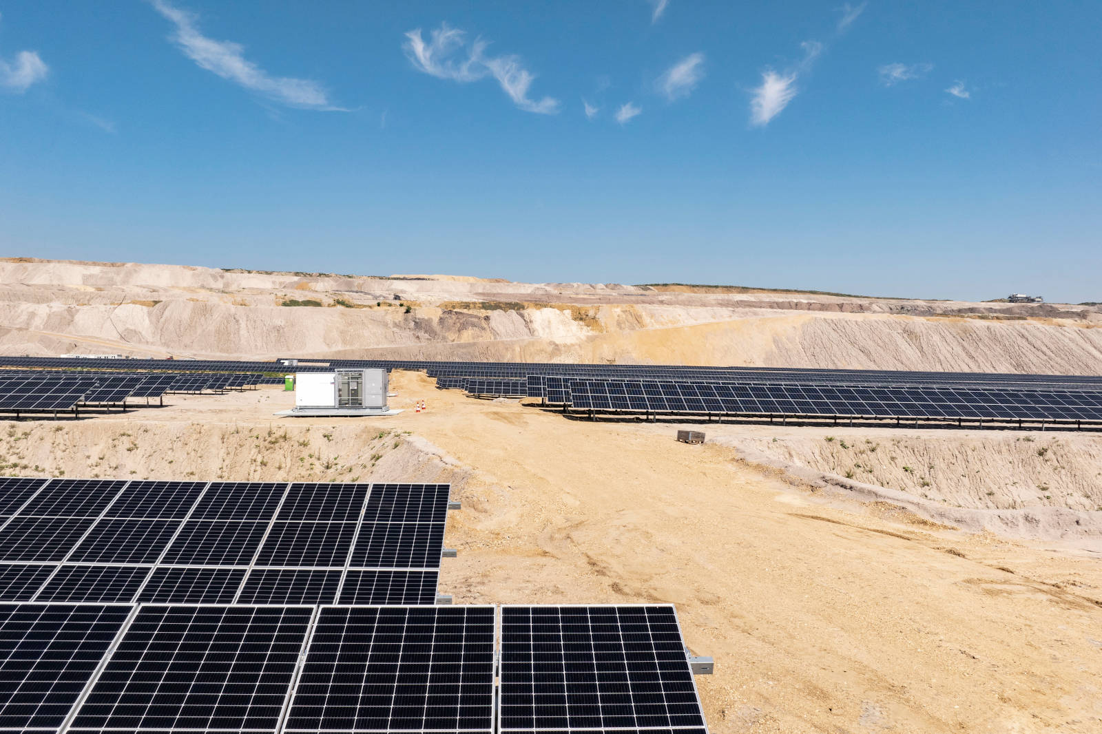 Ein weitläufiges Solarfeld mit Solarpanelen, umgeben von sandigen Flächen und sanften Hügeln unter einem blauen Himmel.