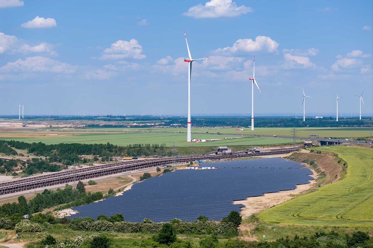 A wide landscape featuring wind turbines, solar panels, and a body of water in the foreground. The sky is blue with some clouds.