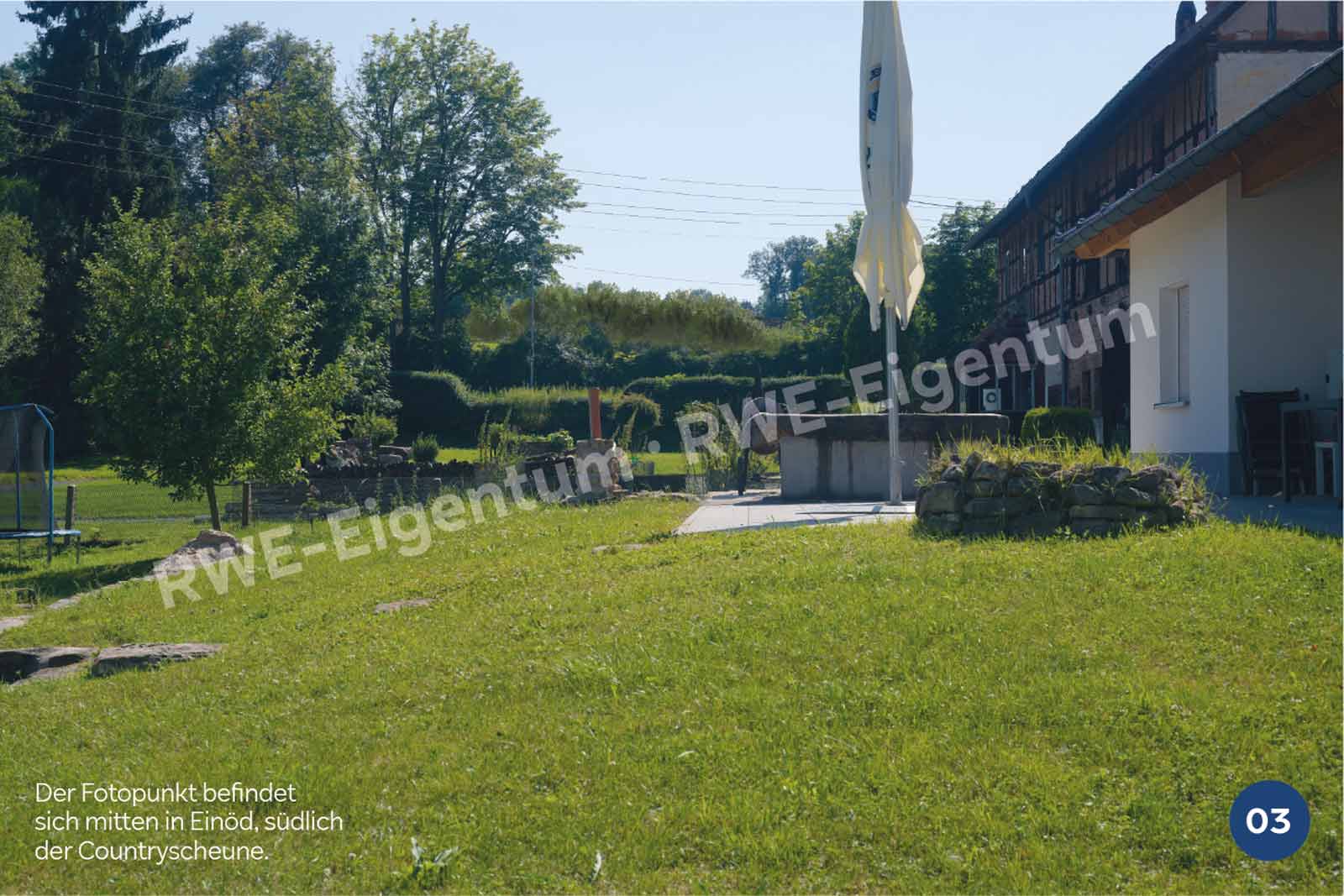 Eine sonnige Gartenszene mit einem Trampolin, grünem Gras, Bäumen und einer Terrasse mit einem Sonnenschirm neben einem rustikalen Gebäude.