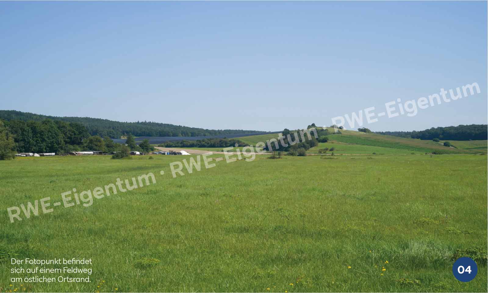 Ein üppig grünes Feld mit entfernten Hügeln und Bäumen unter einem klaren blauen Himmel, vom Rand eines Dorfes aus betrachtet.