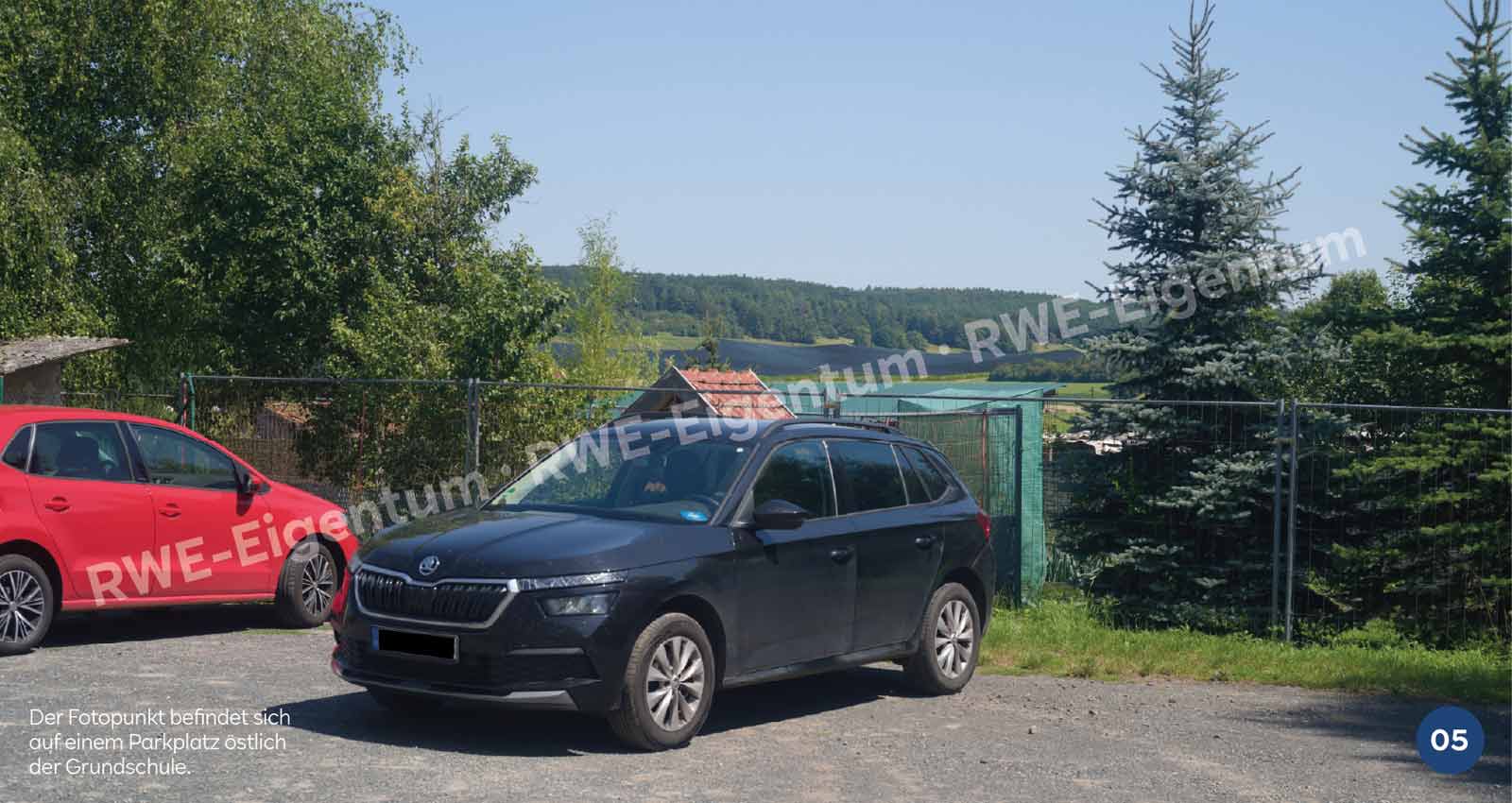 Ein schwarzes Auto steht auf einem Kiesparkplatz neben einem roten Auto, mit einer grünen Landschaft im Hintergrund.