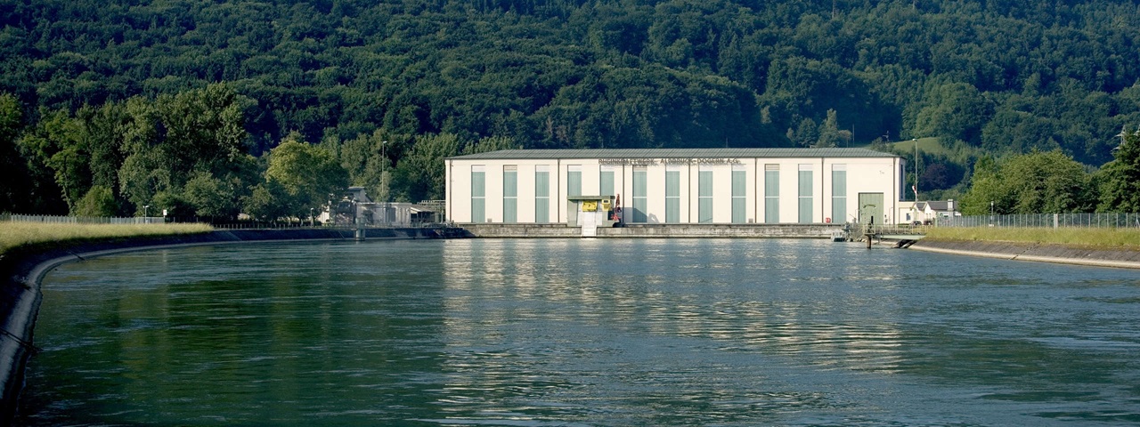 A modern building by a waterway, surrounded by trees and hills, reflected in the calm water.