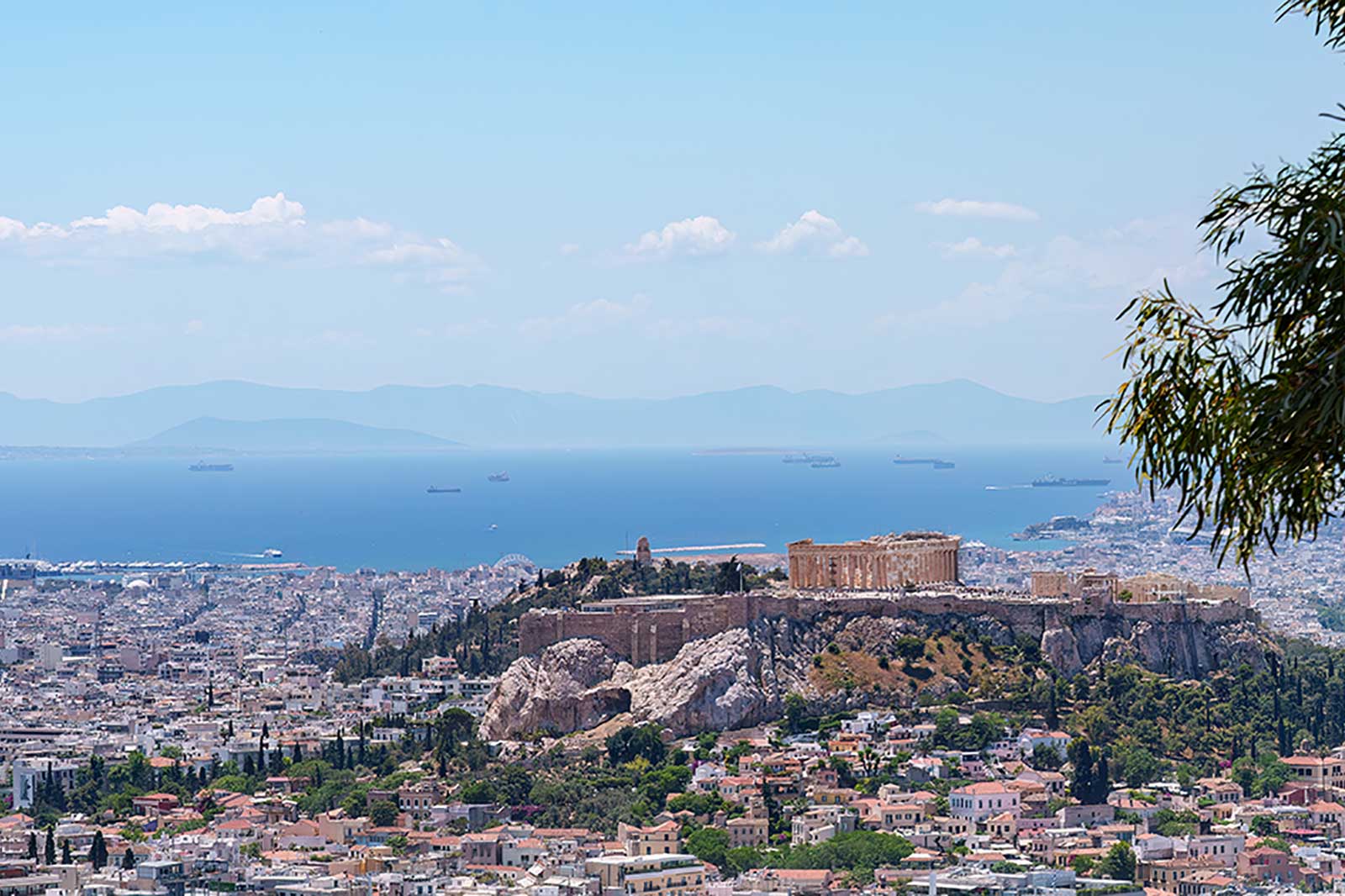 A panoramic view of Athens, showcasing the Acropolis, the sea, and distant mountains under a clear blue sky.
