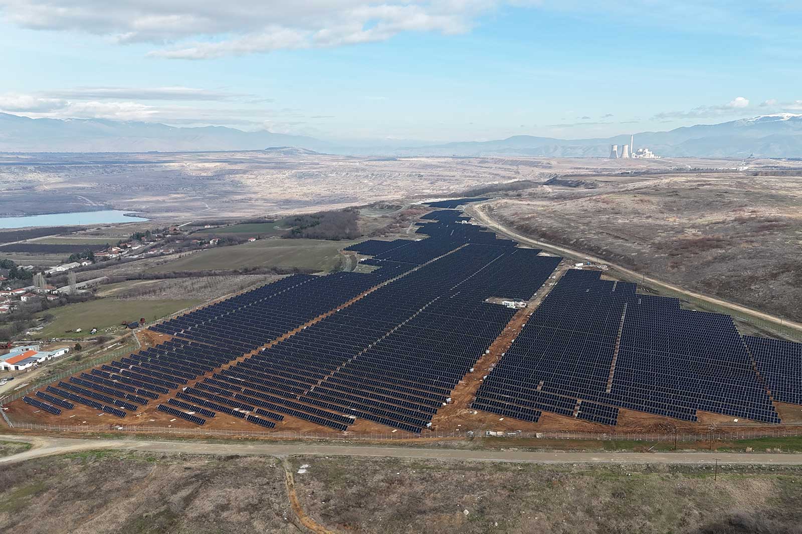 Aerial view of a large solar farm with rows of solar panels, surrounded by hills and a small water body in the distance.