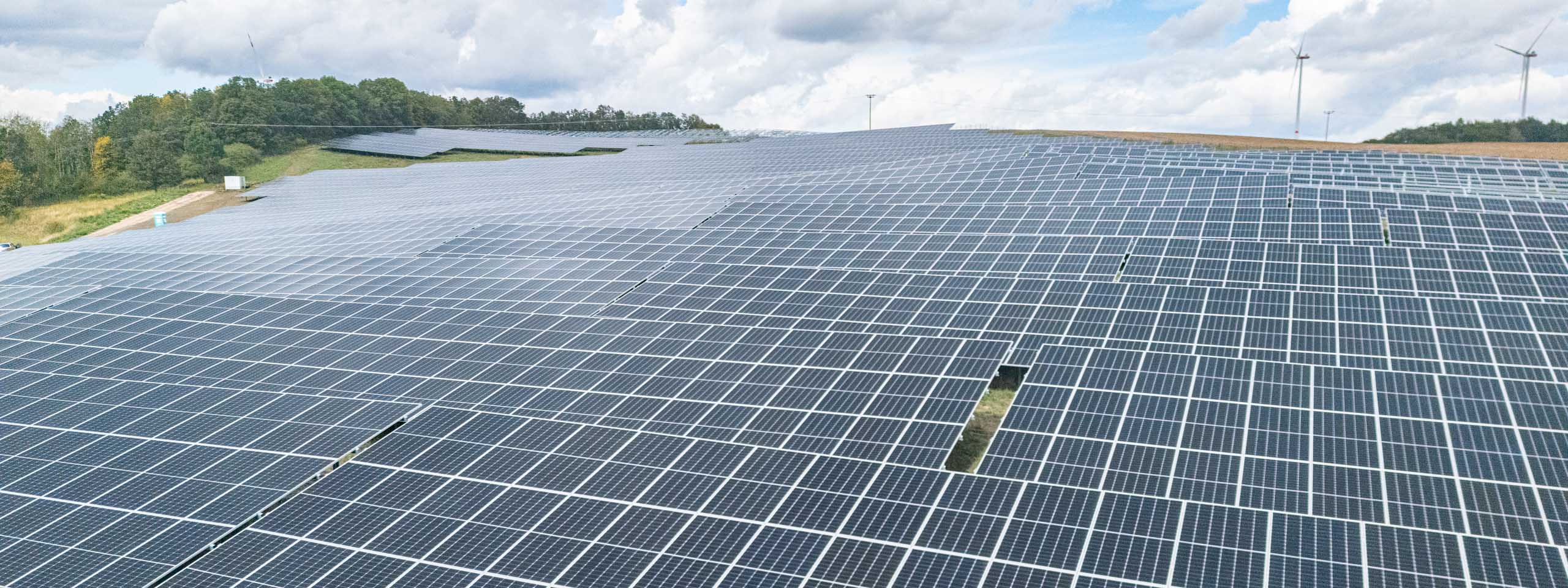 Luftaufnahme eines Solarparks mit Reihen von Solarpanelen auf einem Hügel unter einem teils bewölkten Himmel.
