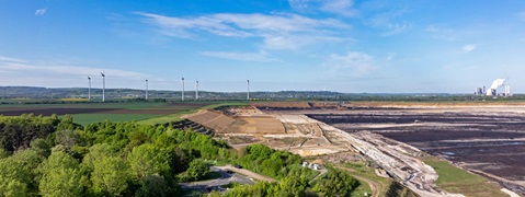A wide landscape featuring wind turbines, adjacent to excavated land and a prominent industrial facility in the background.