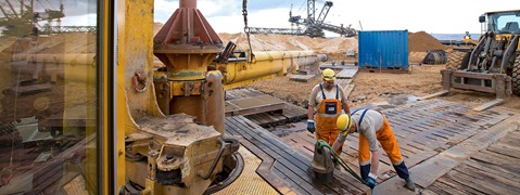 Construction workers operate machinery at a site with heavy equipment and sand in the background.