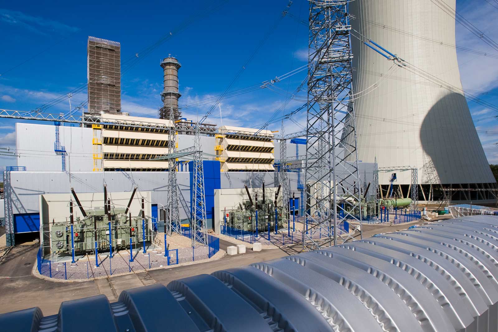 The image shows a power plant with several large transformers and a cooling tower under a clear blue sky.