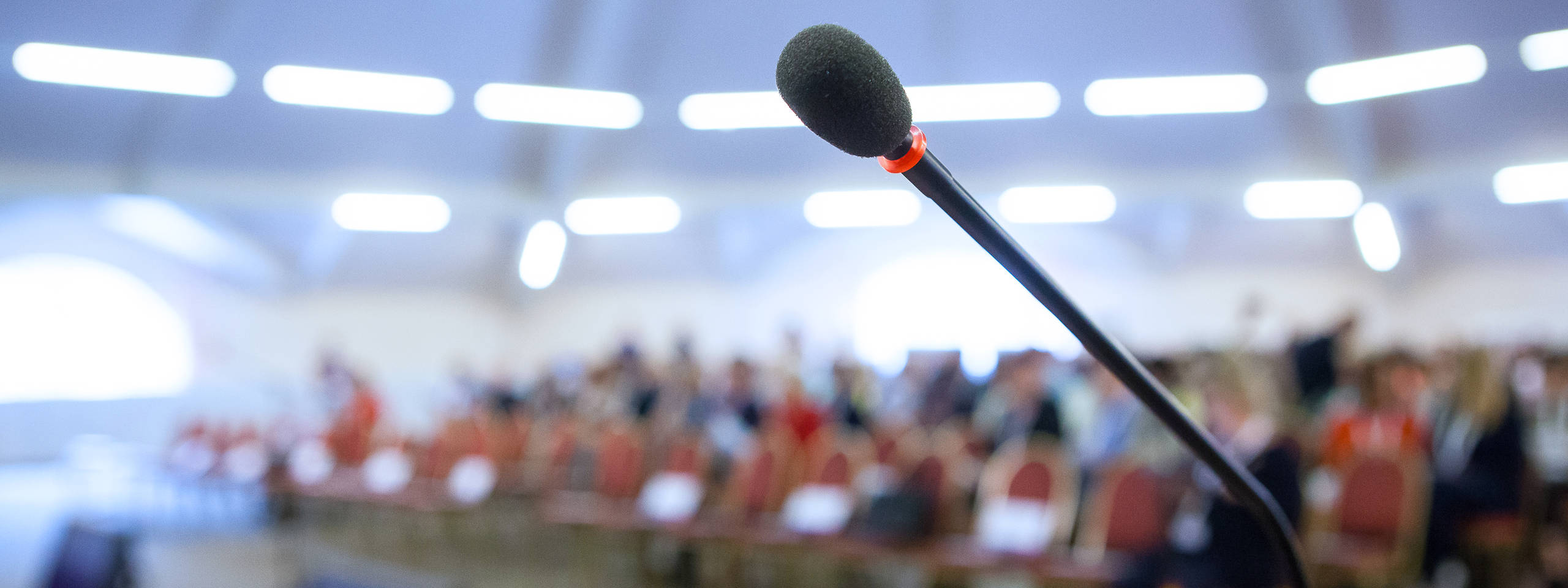 Ein Mikrofon steht im Fokus, während der Hintergrund einen Raum mit sitzenden Personen zeigt.