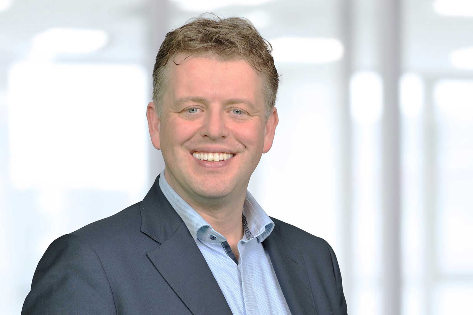 Ein Mann in einem Anzug mit einer blauen Bluse steht vor einem hellen, unscharfen Hintergrund.