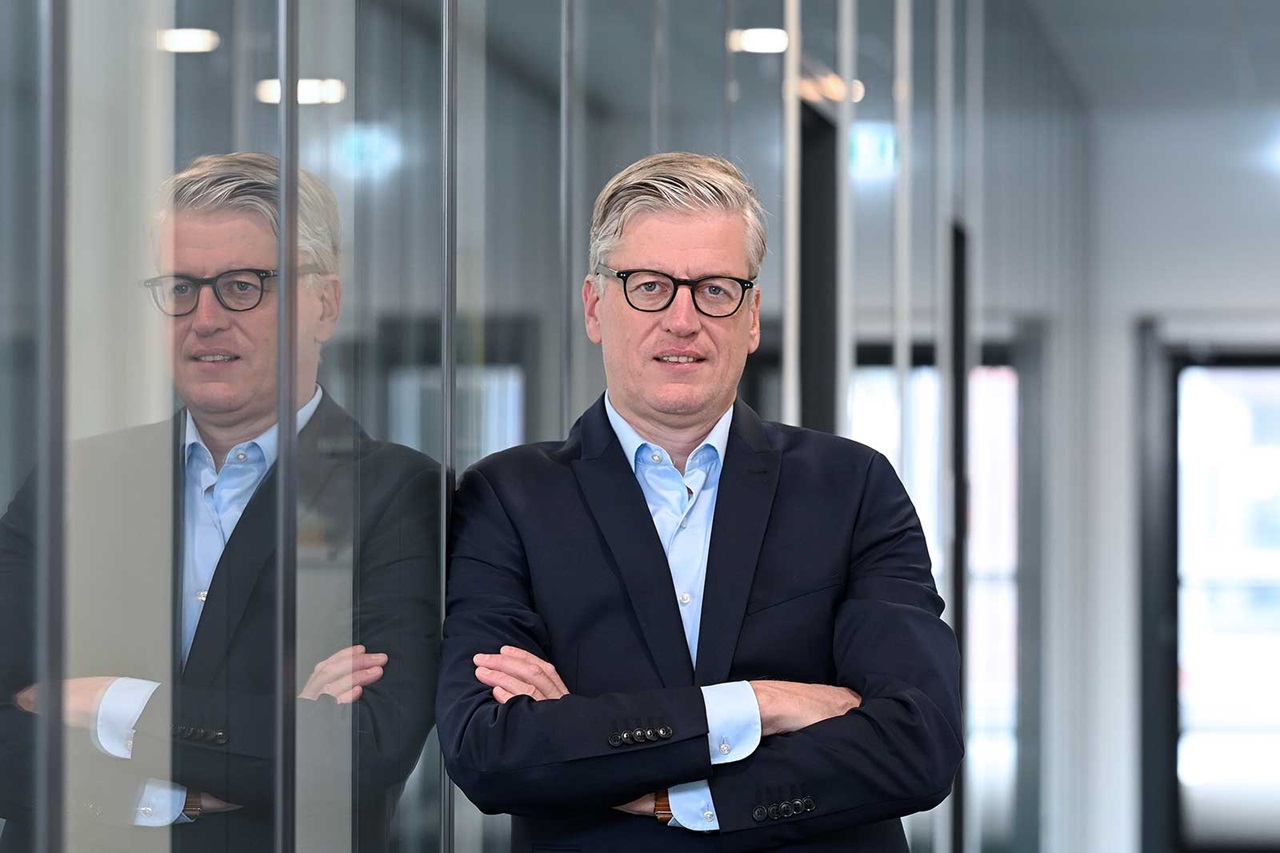 A man in a suit stands casually in front of a glass wall in a modern office.