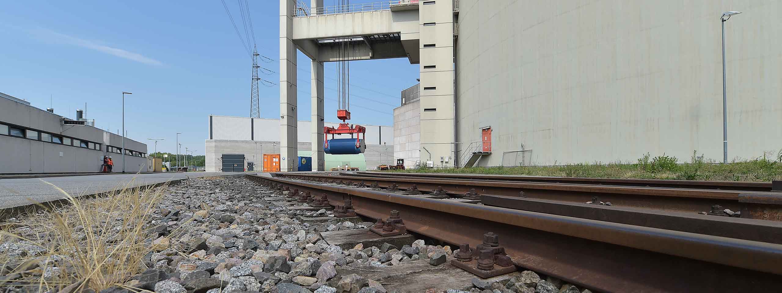 Ein Kran hebt einen blauen zylindrischen Gegenstand in der Nähe von Bahngleisen und Industriegebäuden unter blauem Himmel.