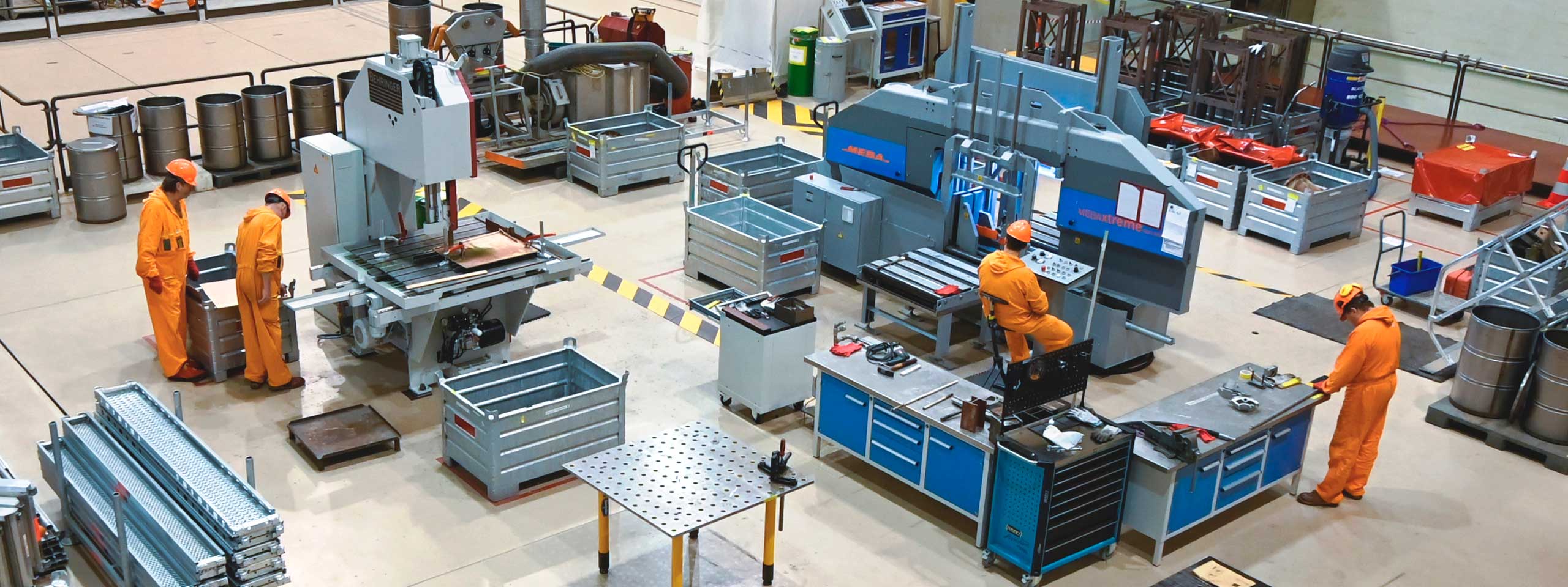 Eine Werkstatt mit mehreren Arbeitern in orangefarbener Arbeitskleidung, Maschinen und Werkzeugen sind sichtbar.