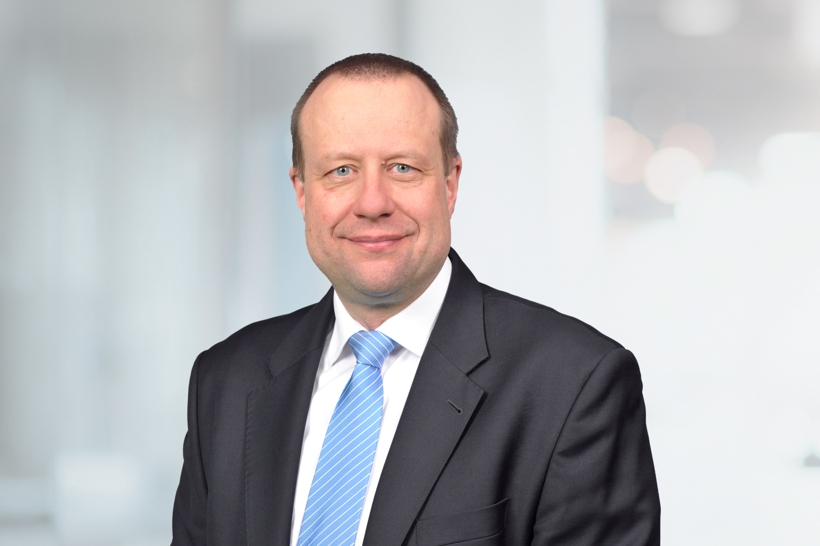 Der Mann trägt einen Anzug mit einer weißen Bluse und steht vor einem hellen Hintergrund.