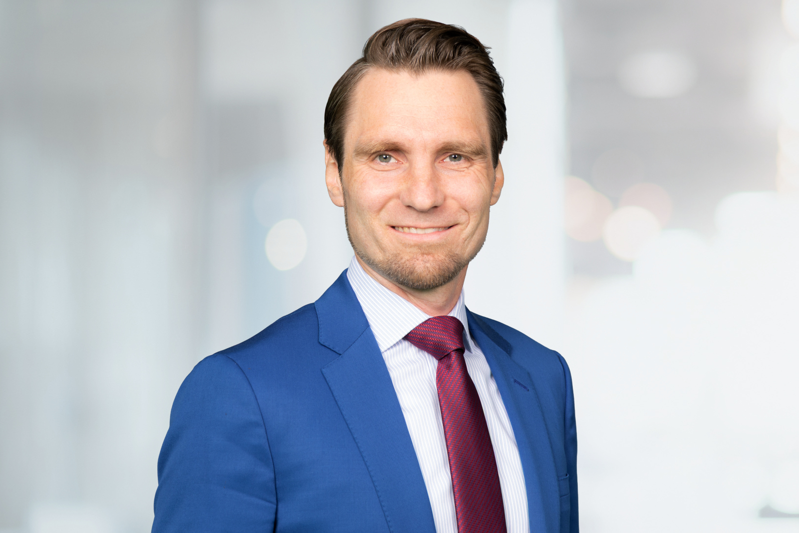 Ein Mann in einem blauen Anzug mit einer roten Krawatte steht vor einem neutralen grauen Hintergrund.