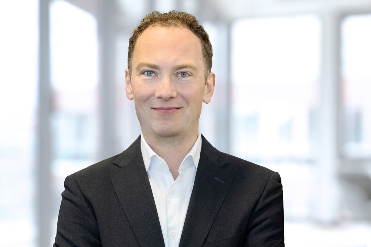 Ein professioneller Mann in einem schwarzen Anzug und einem weißen Hemd steht mit verschränkten Armen in einer hellen, modernen Büroumgebung. Portrait von Dr. Tobias Keitel.