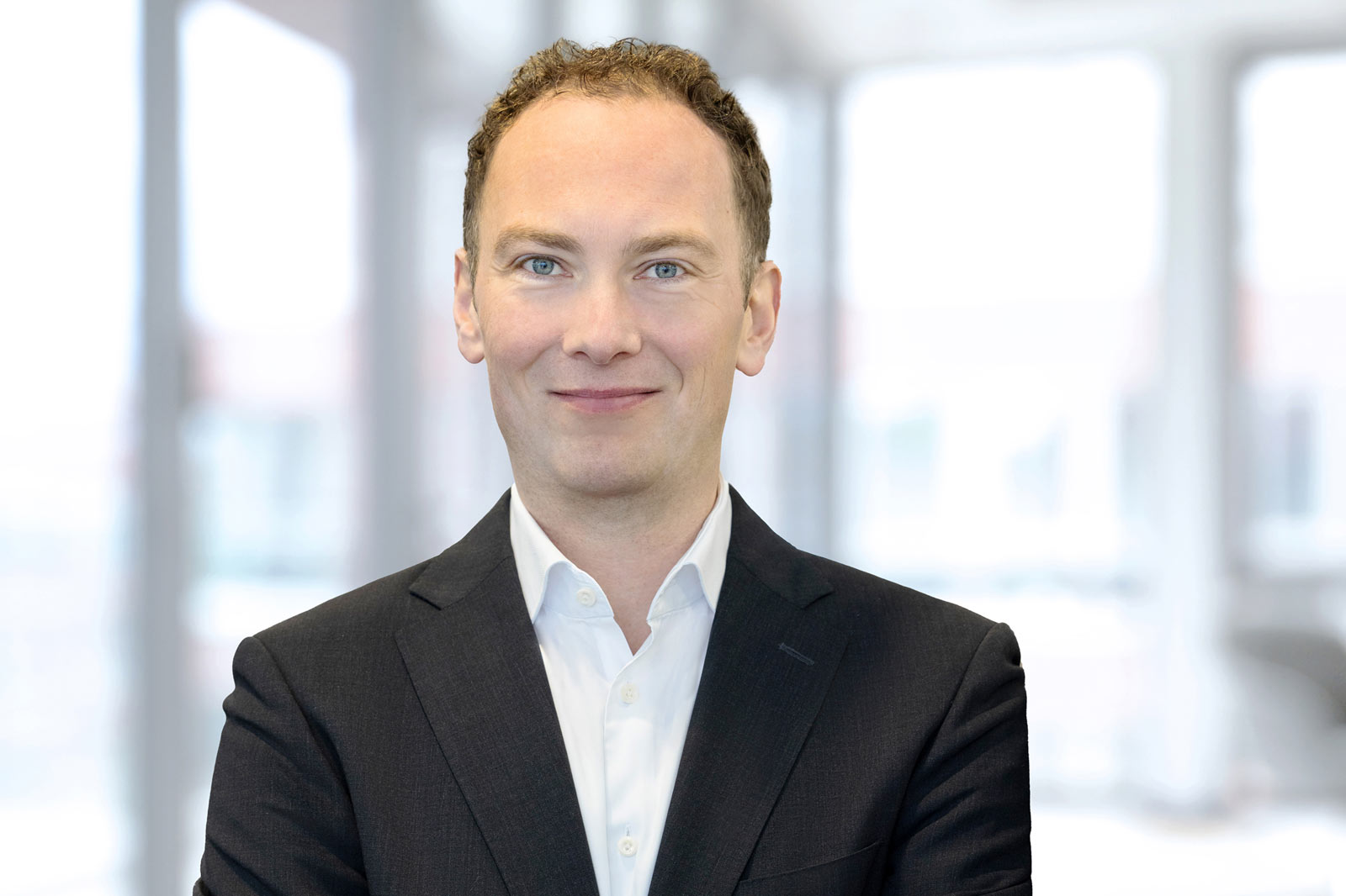 A professional man in a black suit and white shirt stands with arms crossed in a bright, modern office environment. Portrait of Dr. Tobias Keitel.
