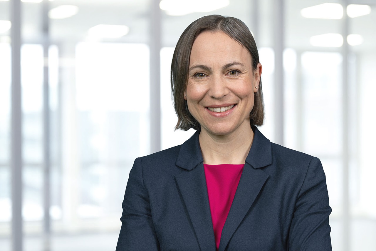 A woman with short dark hair wearing a navy blazer and magenta blouse stands with arms crossed in a bright office with large windows. Portrait of Sandra Dettmer.