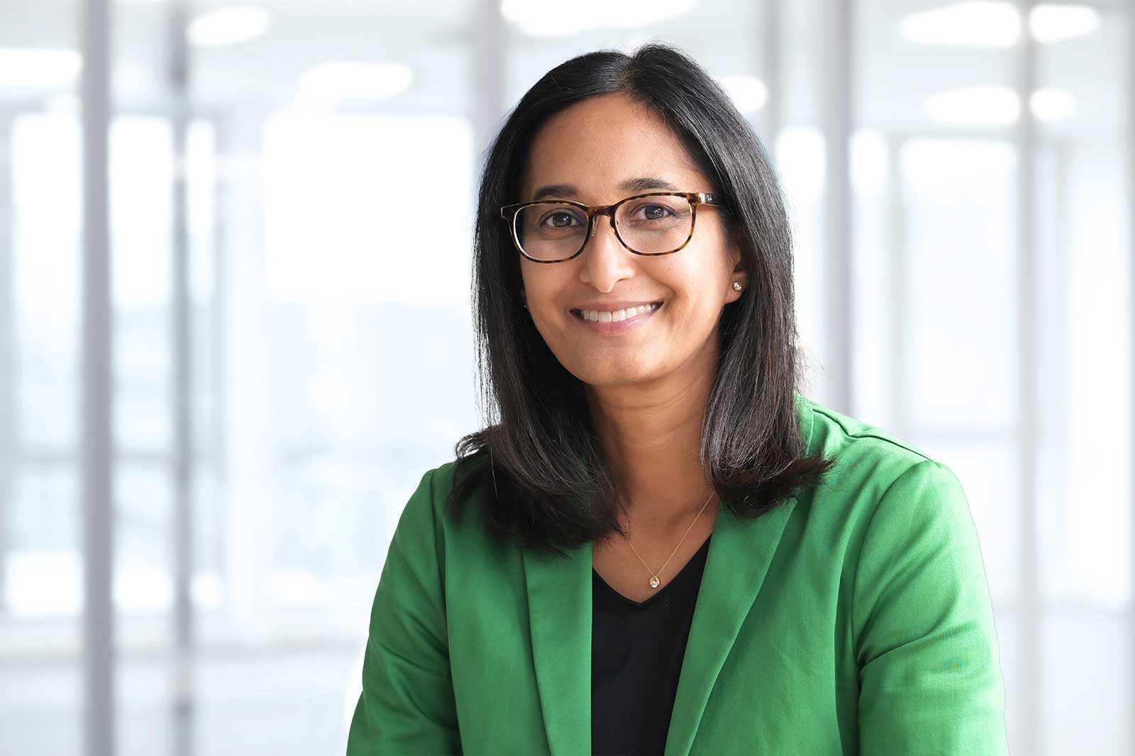 A person with straight black hair, wearing a green blazer and a black top, is sitting in an office environment. Portrait of Sopna Sury.