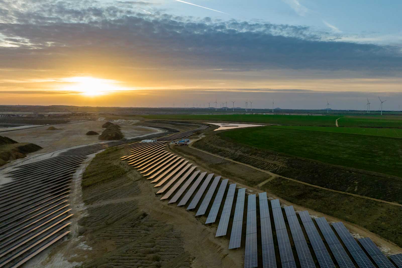 Wide-angle view of a solar power plant at sunset, with wind turbines visible in the background.