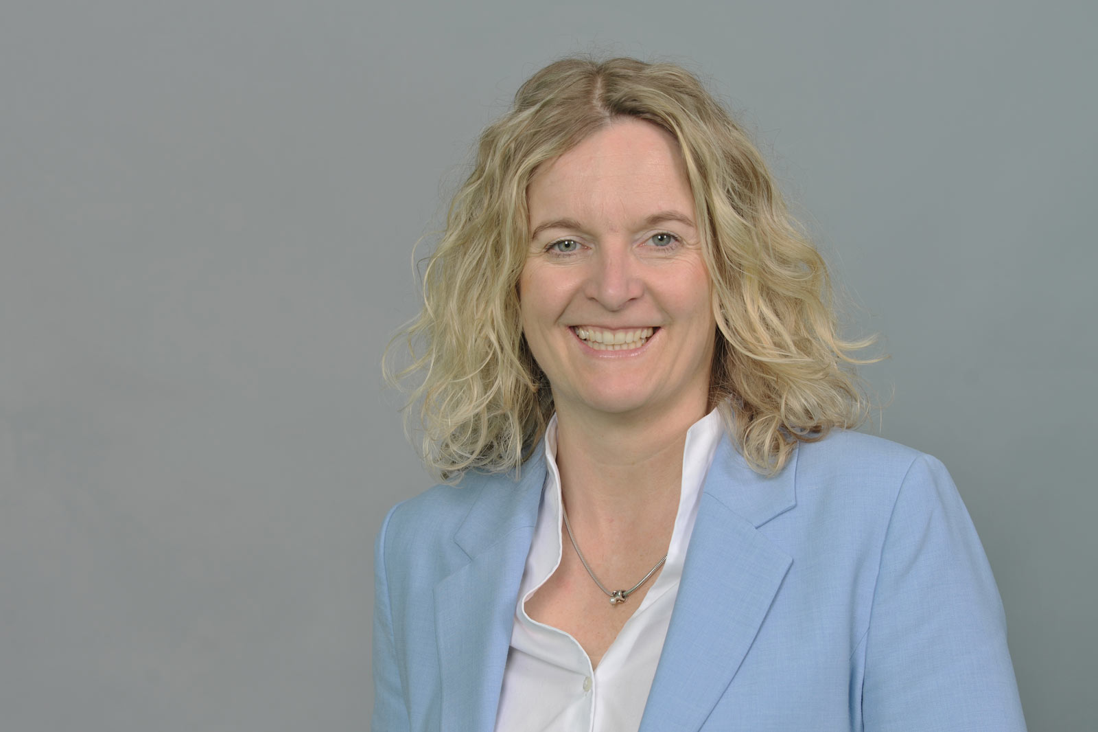A person with curly blonde hair is wearing a light blue suit and a white shirt against a grey background. Portrait of Anja Dubbert.