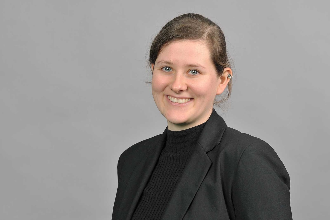 Eine Person in einem schwarzen Blazer und Rollkragenpullover vor einem grauen Hintergrund. Portrait von Anne Fricke.