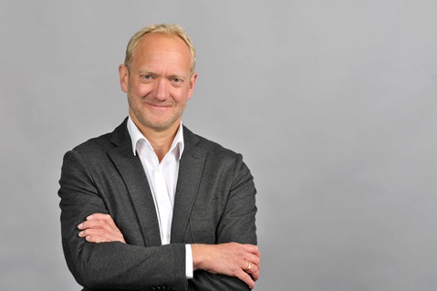 A person in a grey suit and white shirt stands with crossed arms in front of a grey background. Portrait of Ansgar Vierhaus.