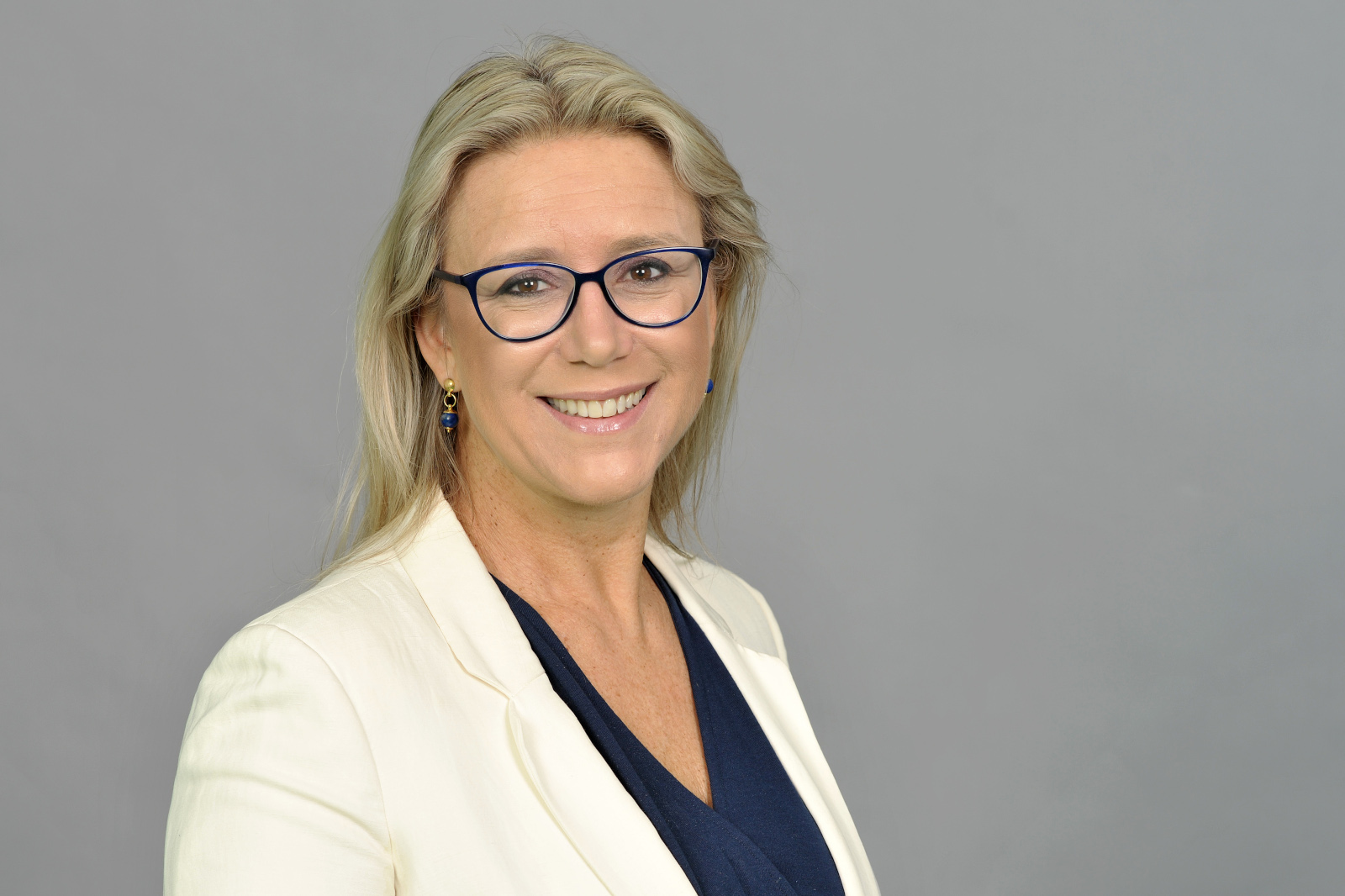 A woman with long, blonde hair wears a blazer over a navy top against a grey background. Portrait of Federica Maranca.