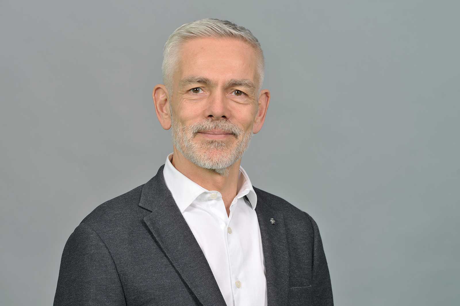 A man in a grey suit and white shirt stands against a grey background. Portrait of Gareth Cribb.