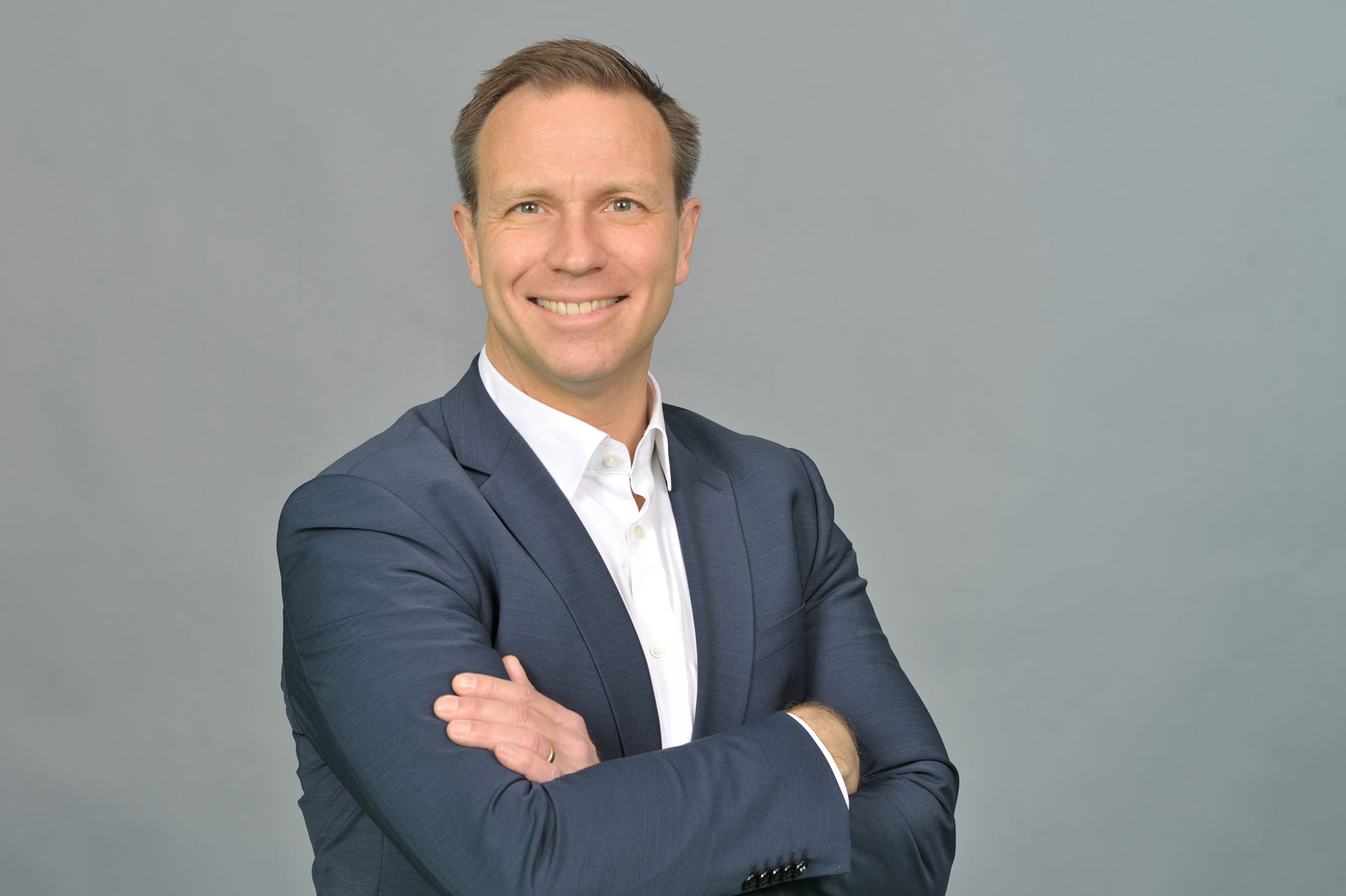 A man in a dark suit with crossed arms, standing against a grey background. He is wearing a white shirt. Portrait of Götz Hanau.