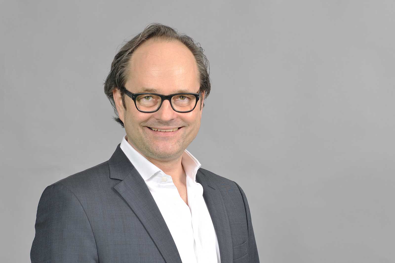 A person wearing a dark suit and white shirt poses against a plain grey background. Portrait of Henning Joswig.