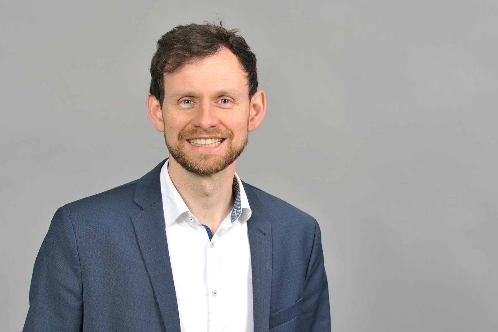 A man is wearing a suit and a shirt, standing against a grey background. Portrait of Jan Josef Bernholz.
