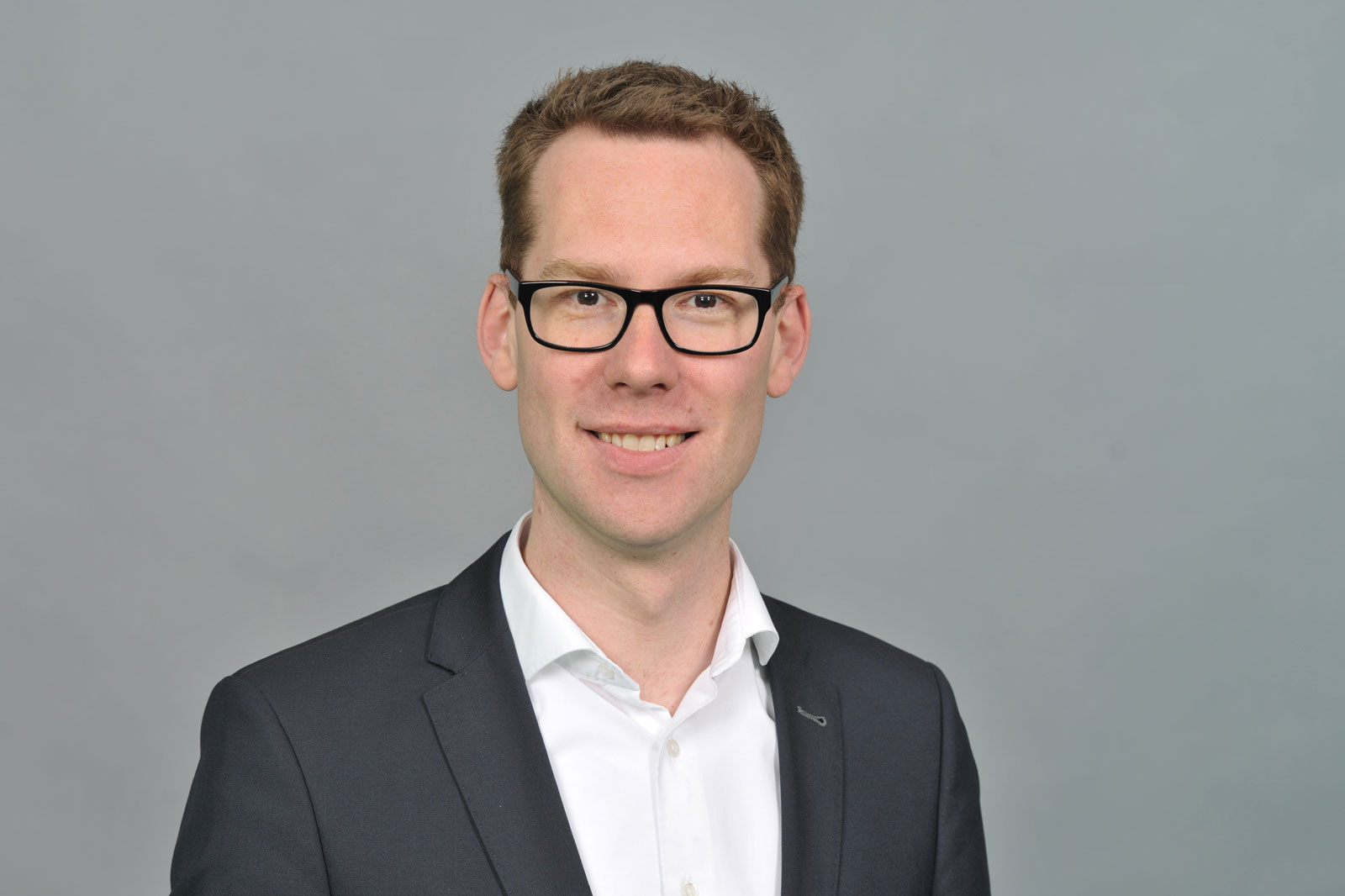A man is wearing a black suit and a white shirt. The background is grey. Portrait of Kevin Pasch.