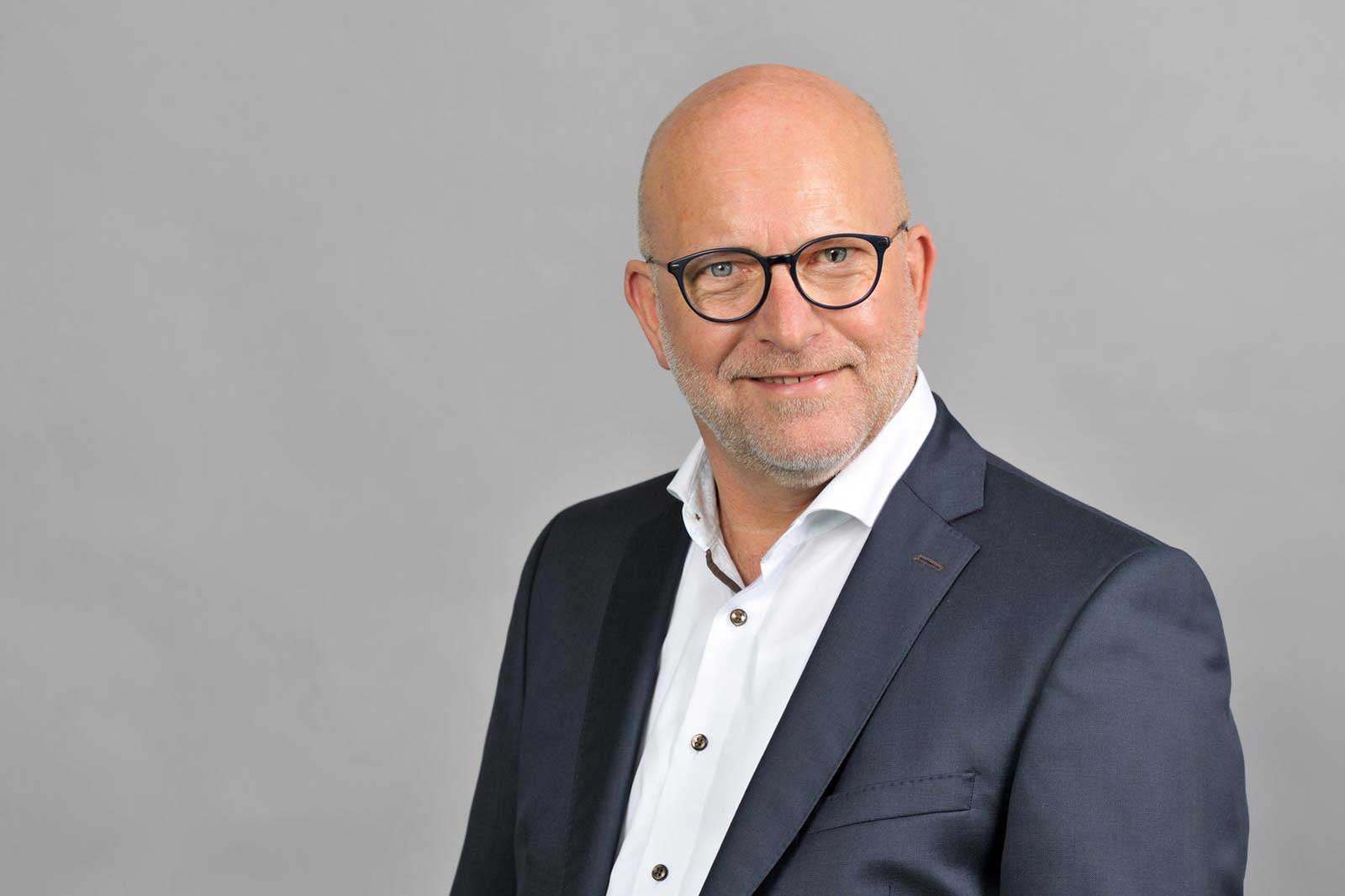 A man in a formal suit stands against a grey background, his facial features are obscured. Portrait of Michael Schütz.